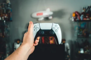 Hand holding a white PlayStation DualSense controller in a gaming room